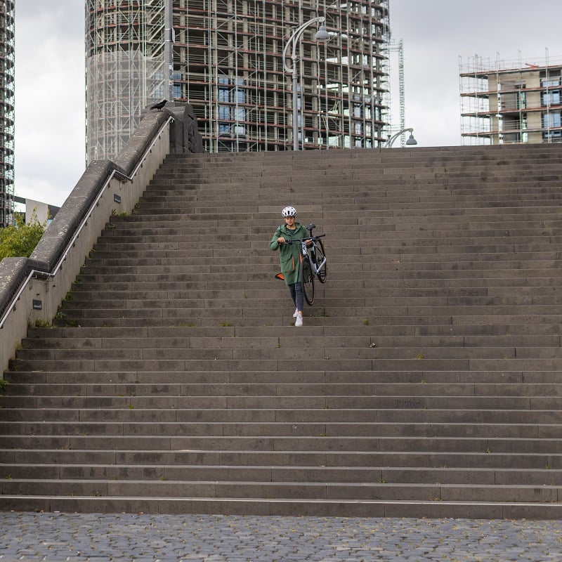 Person mit Fahrrad und Helm geht große Treppe hinunter, Baustellengebäude im Hintergrund.