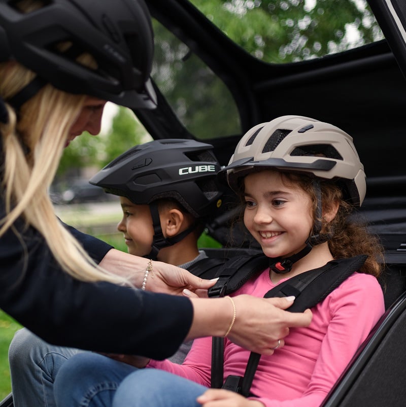 Zwei Kinder mit Helmen sitzen im Cube Trike Hybrid, ein Erwachsener schnallt Sicherheitsgurte an.
