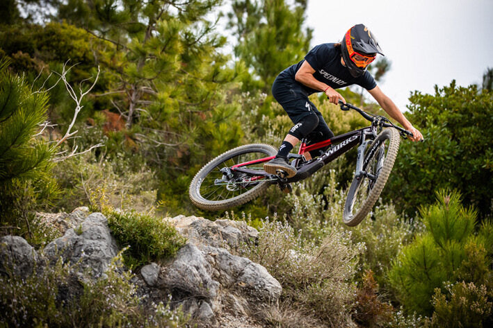 Mountainbiker auf Specialized Bike springt über Felsen im Wald. Ridewear und Helm zeigen Fokus auf Offroad-Action.