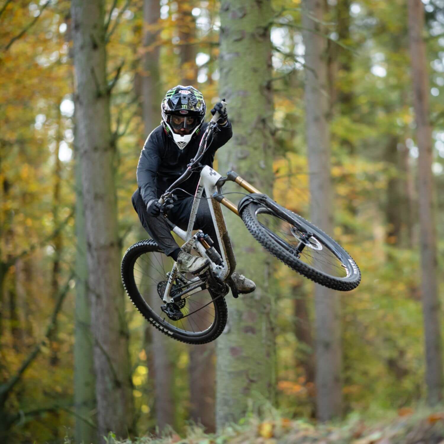 Mountainbiker springt mit Downhill-Bike durch Wald, trägt Schutzkleidung und Helm. Ideal für actionbegeisterte Outdoor-Fans.