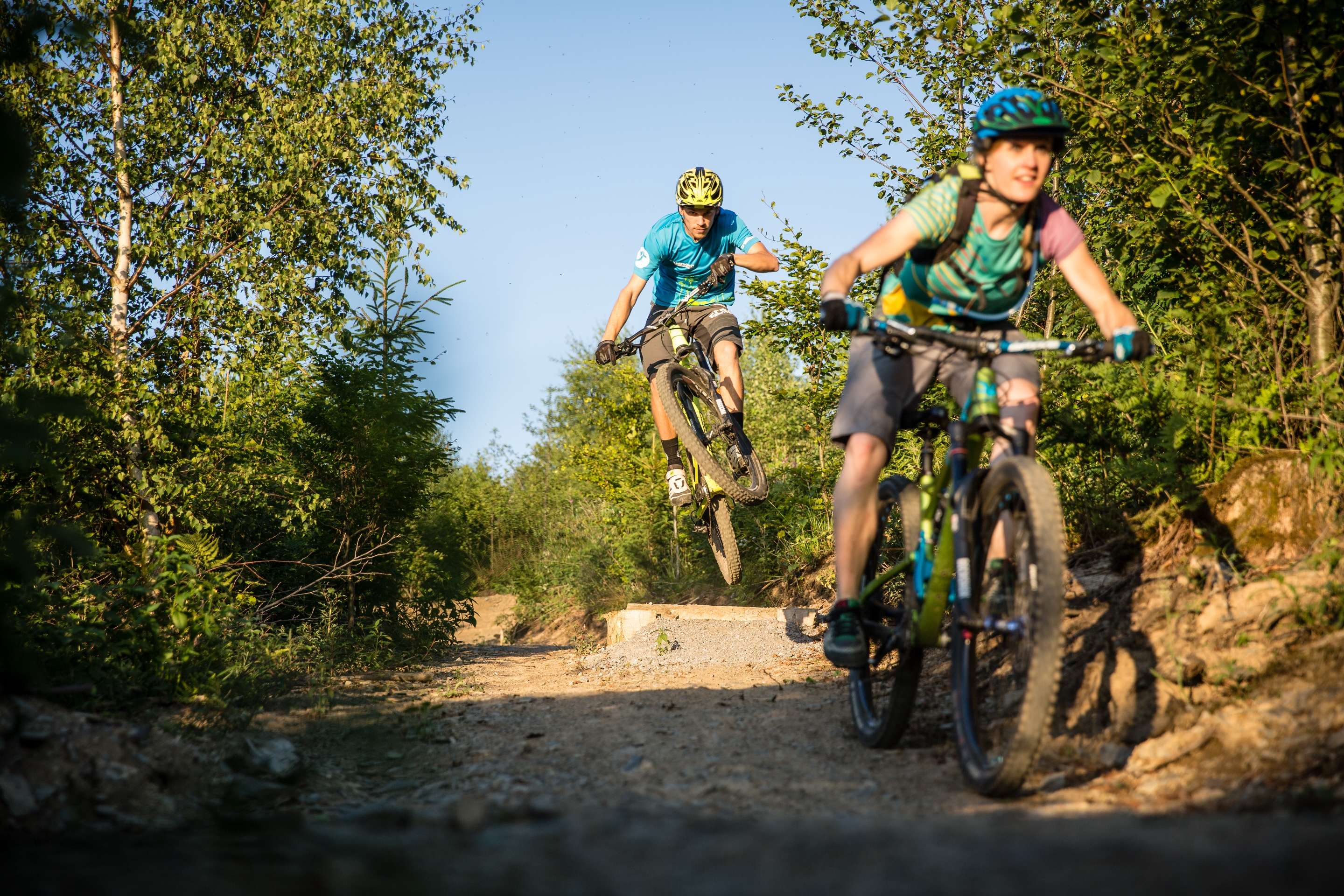 Zwei Mountainbiker mit Helmen fahren auf einem Waldweg, einer macht einen Sprung, sommerliche Umgebung.