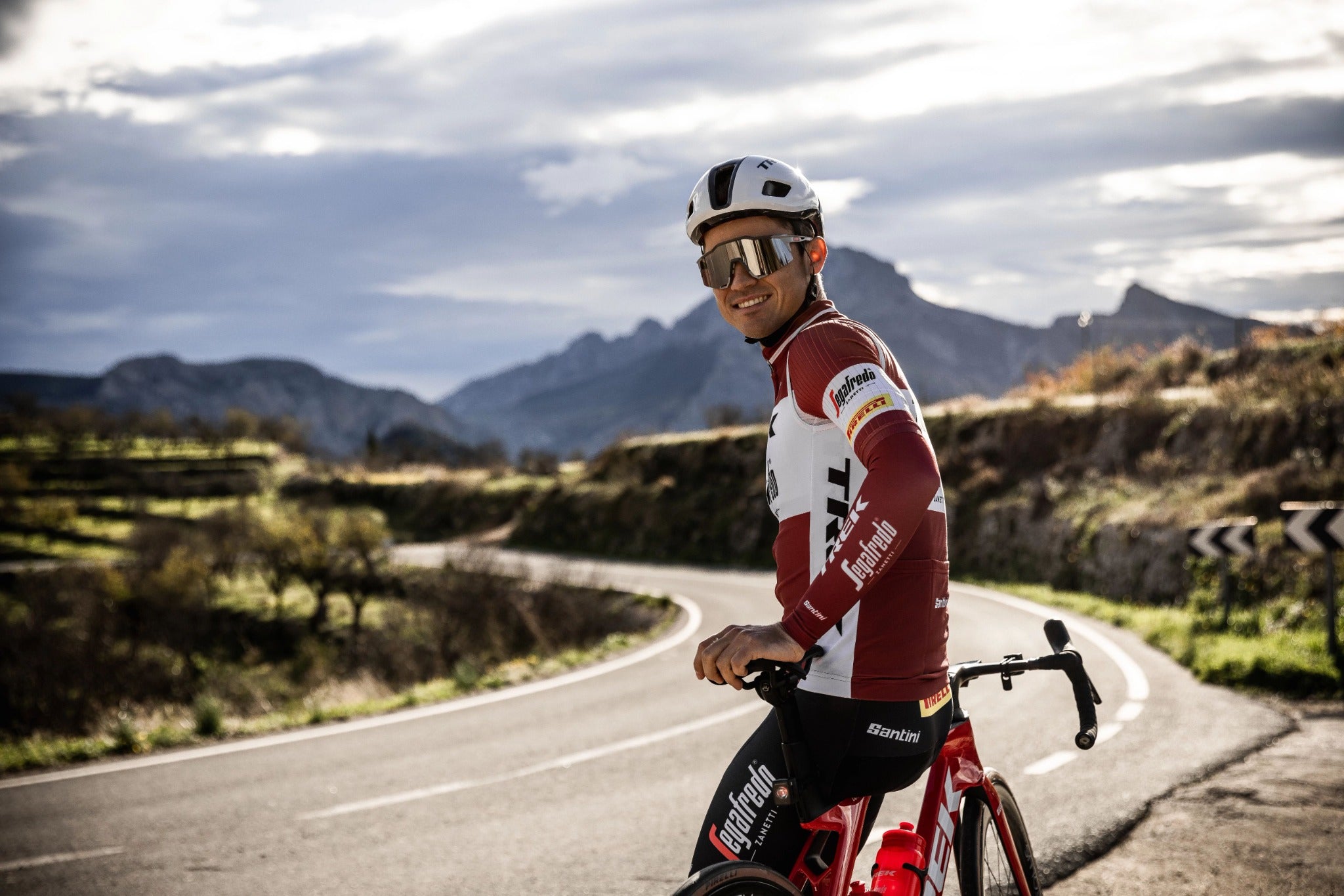 Radfahrer mit Trek-Fahrrad und Trek-Segafredo-Kleidung auf kurviger Landstraße, Berge im Hintergrund.