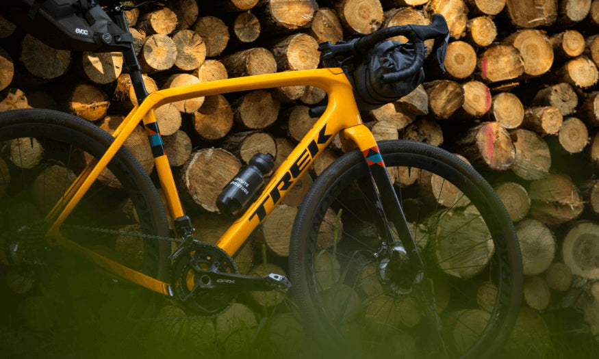 Gelbes Trek Gravelbike neben Holzstapeln, ausgestattet mit Trinkflasche und Lenkertasche. Ideal für Outdoor-Abenteuer.