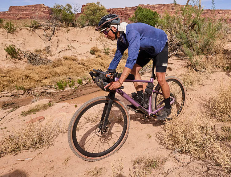 Radfahrer in blauer Kleidung auf einem lilafarbenen Gravelbike in sandiger Wüstenlandschaft, geeignet für Offroad-Abenteuer.