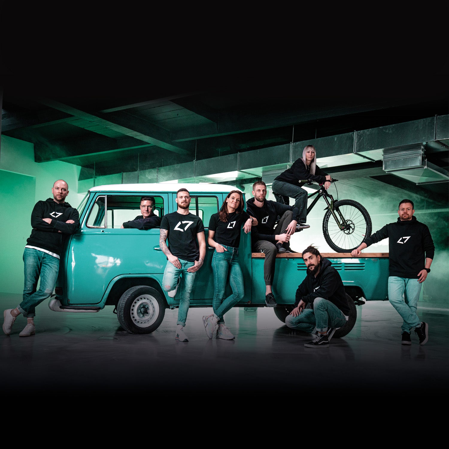 Gruppe von Menschen in Freizeitkleidung posiert vor einem blauen Transporter mit Fahrrad auf dem Dach.