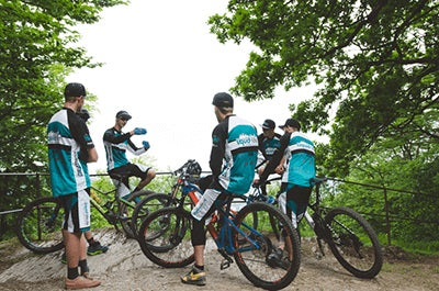 Gruppe von Mountainbikern in blauer Sportkleidung steht mit Fahrrädern im Wald und bereitet sich auf eine Fahrt vor.