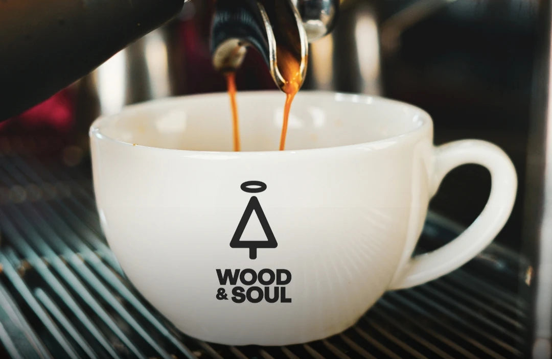 Weiße Kaffeetasse mit "Wood & Soul" Logo, in die Kaffee aus einer Espressomaschine fließt. Ideal für Kaffeeliebhaber.