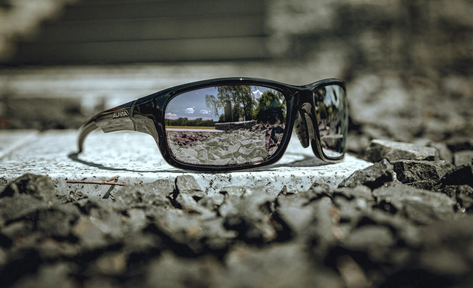 Sonnenbrille von Alpina auf einem steinigen Untergrund, spiegelt eine Landschaft mit Bäumen und blauem Himmel wider.