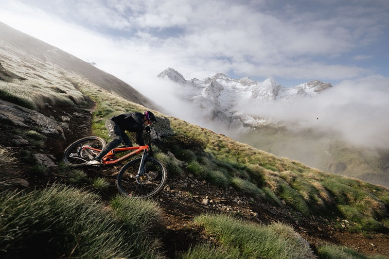 Mountainbiker fährt Santa Cruz Mountainbike auf steilem Bergpfad, schneebedeckte Gipfel im Hintergrund, geeigneter Einsatzbereich: Offroad.