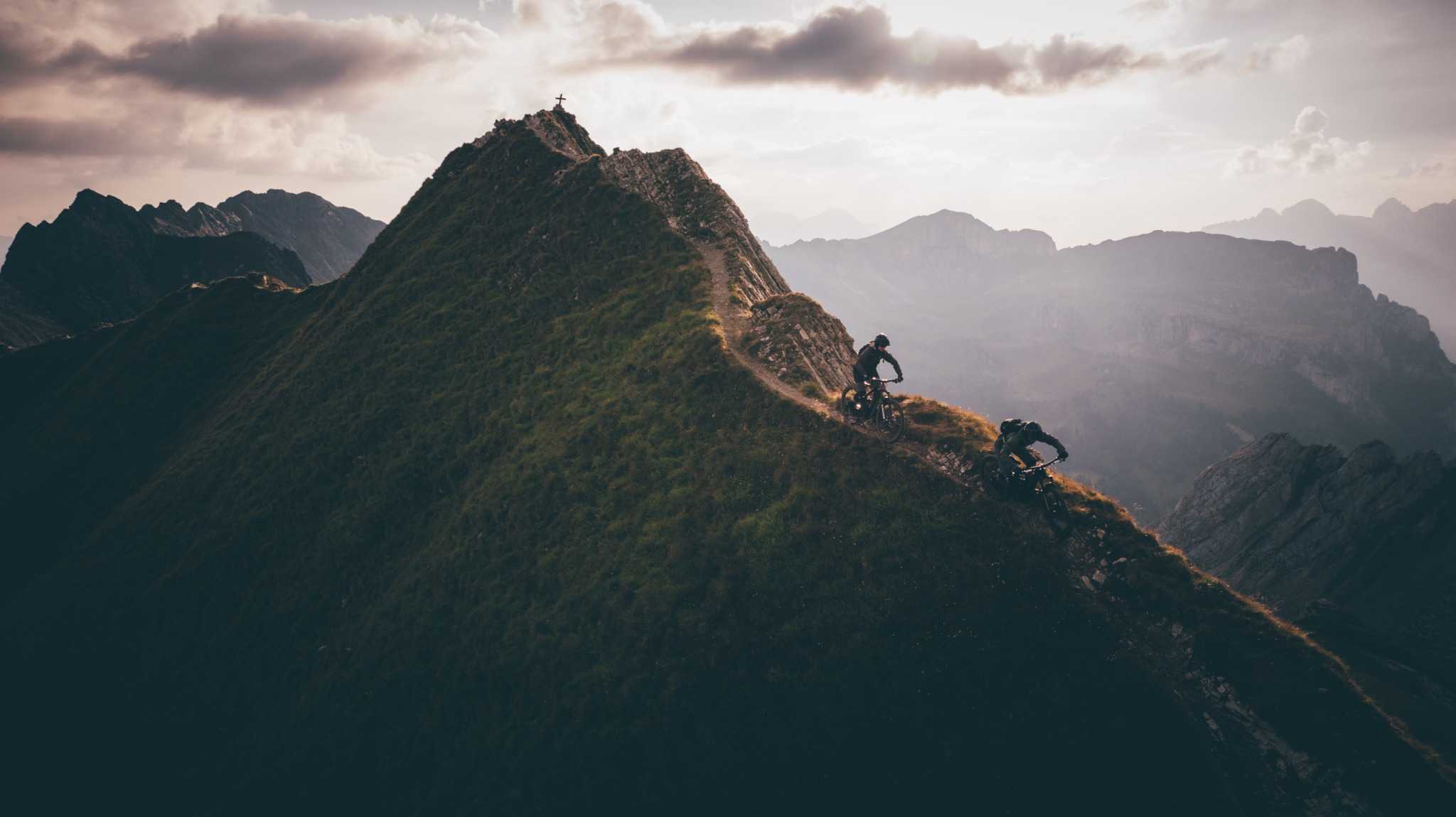 Zwei Mountainbiker fahren auf einem schmalen Bergrückenpfad bei Sonnenuntergang. Abenteuerliche Gebirgskulisse.