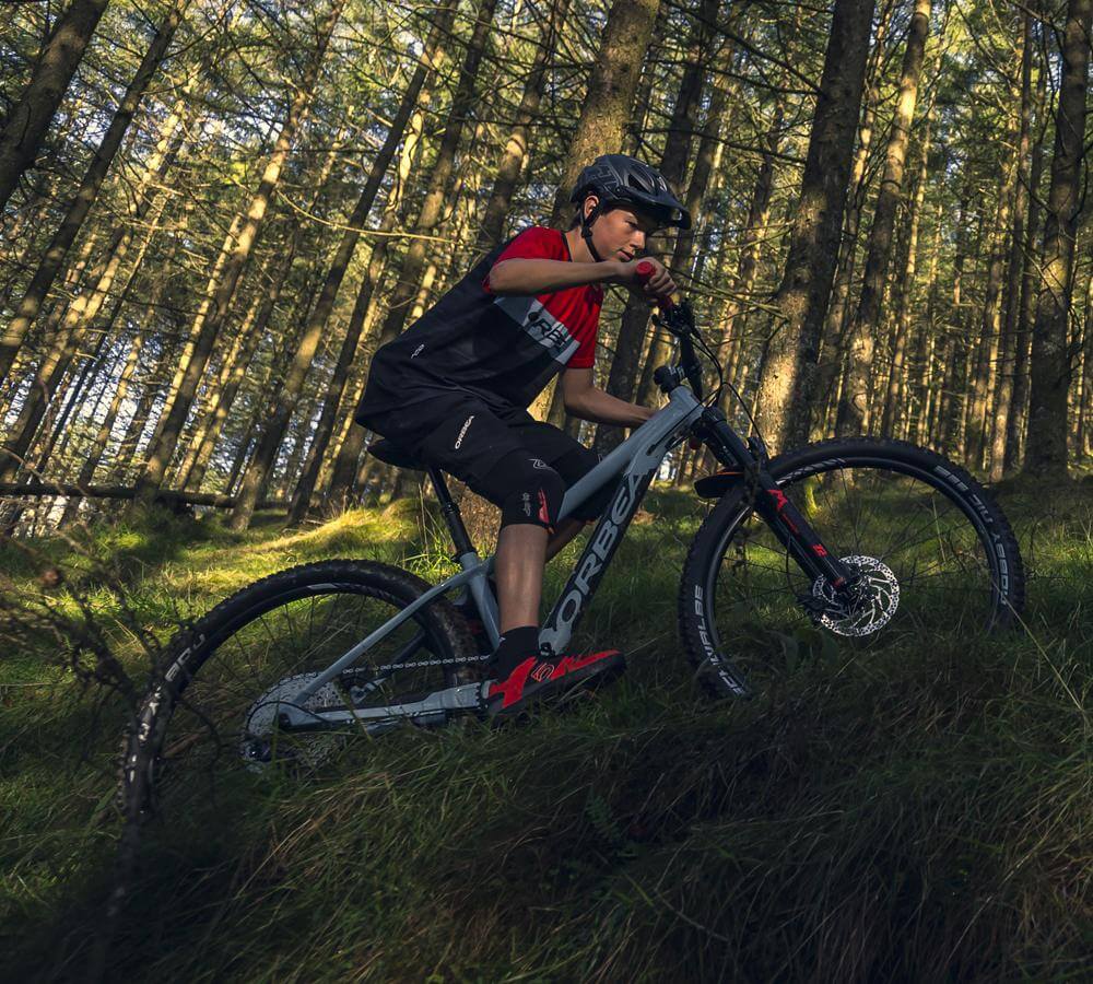 Jugendlicher fährt im Wald ein Orbea Laufey Junior Mountainbike bergauf.
