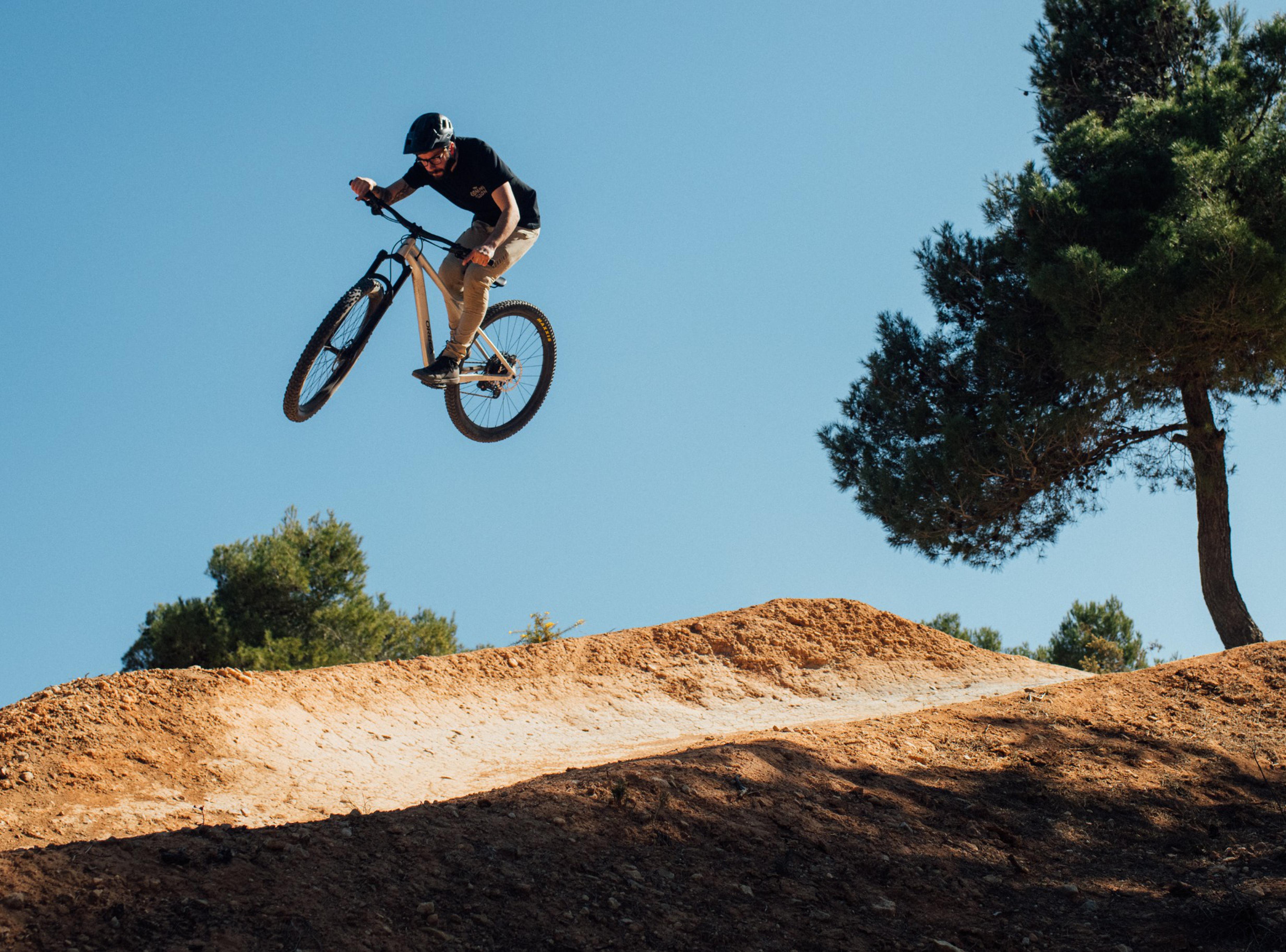 Person auf einem Orbea Laufey Mountainbike macht Sprung auf einem Hügel, sonniger Tag, Wald im Hintergrund.