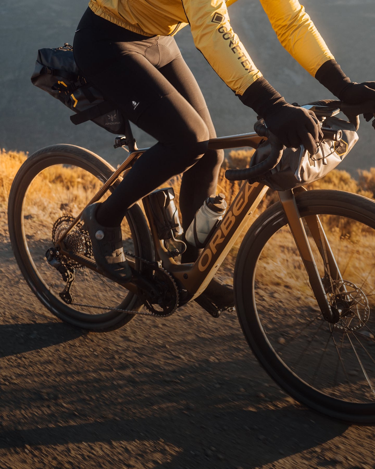 Fahrer auf einem Orbea-Fahrrad mit Tasche, gekleidet in gelber Jacke und schwarzer Hose, auf einer Straße in der Natur.