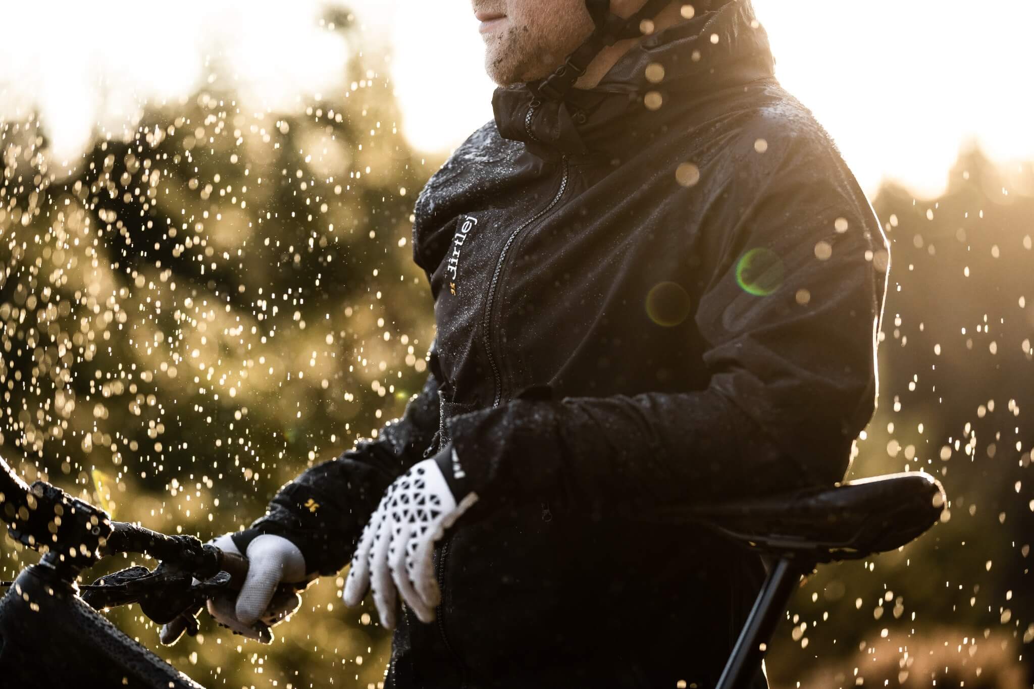 Person trägt Dirtlej-Jacke beim Radfahren im Regen, Sonnenlicht im Hintergrund.