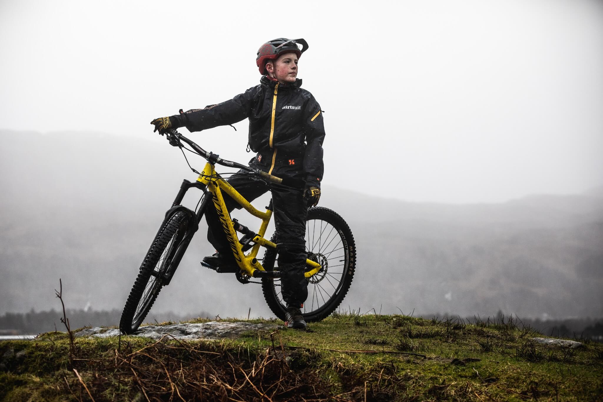 Kind in Fahrradkleidung mit gelbem Mountainbike auf Hügel in nebliger Landschaft.