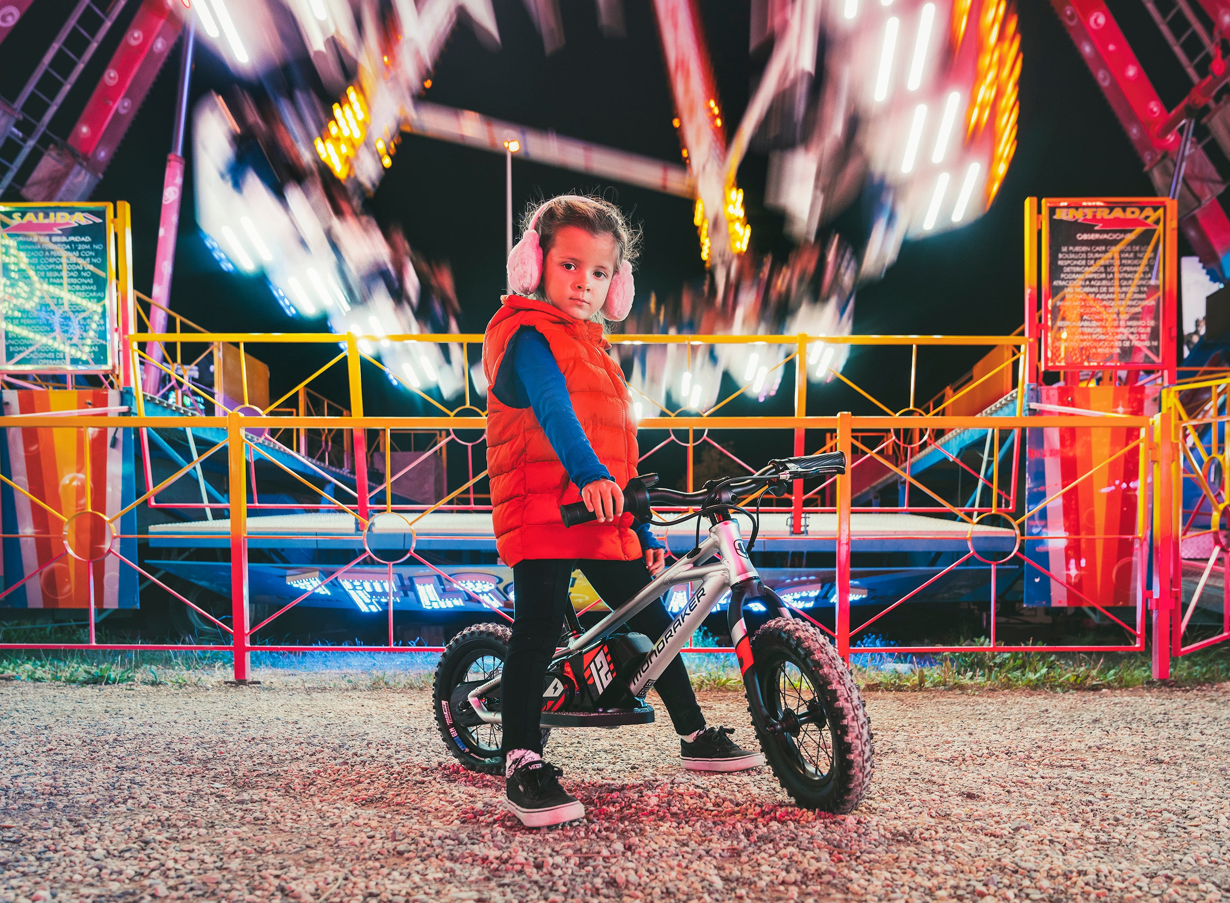 Kind mit Ohrenschützern auf Mondraker-Kinderfahrrad vor beleuchteter Kirmesattraktion bei Nacht.