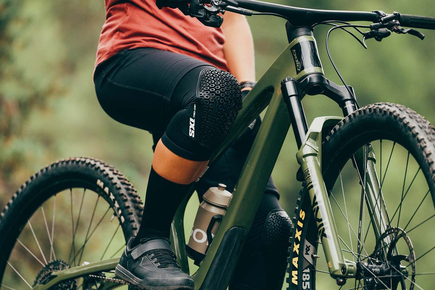 Person auf grünem Mountainbike mit Schutzkleidung, Fokus auf Knieprotektor und Reifen im Waldgelände.