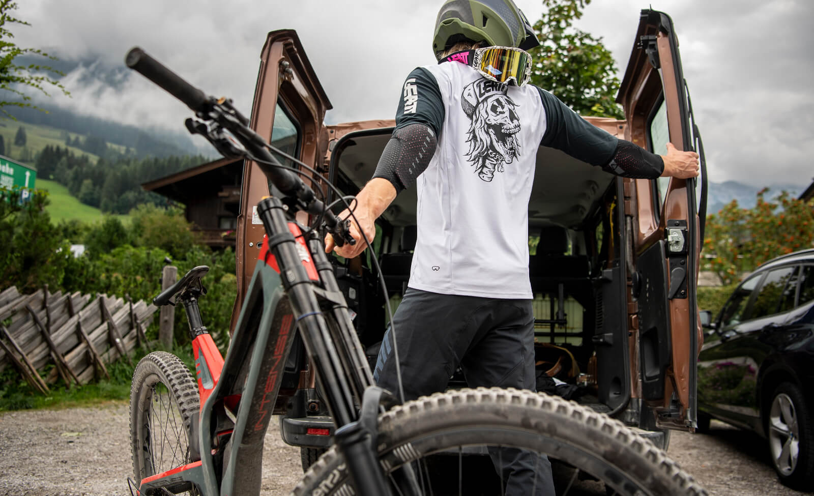 Biker im Leatt-Trikot mit Helm und Brille steht neben einem Mountainbike vor einem geöffneten Van in bergiger Landschaft.
