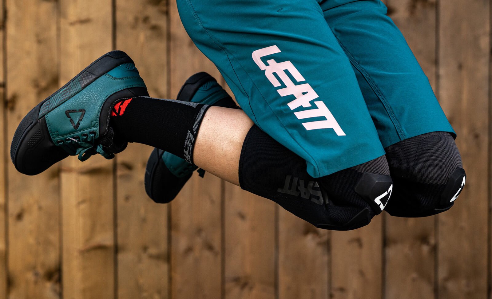 Mountainbike-Ausrüstung von Leatt: Grüne Schuhe, blaue Shorts mit Logo, schwarze Knieschoner in Actionpose.