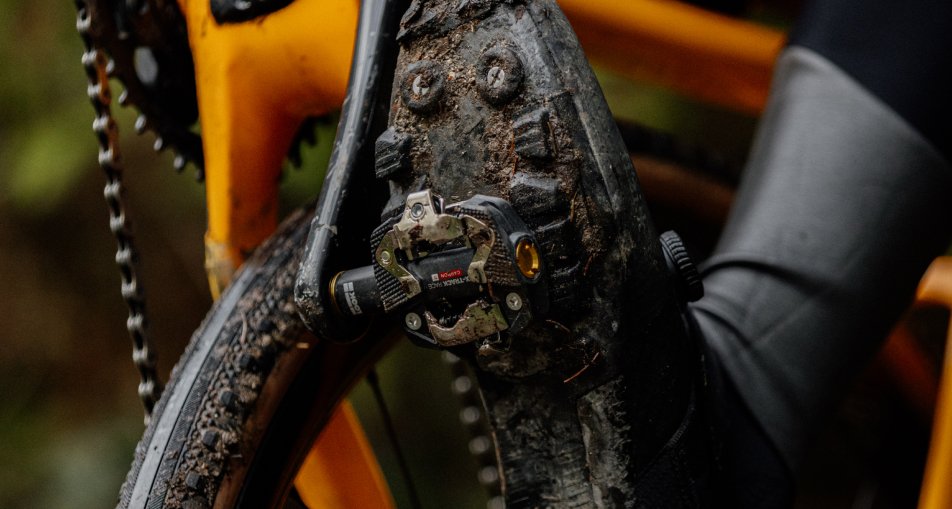 Schuh mit SPD Mountainbike-Pedal, verschmutzt von Matsch, an einem orangefarbenen Fahrrad montiert.