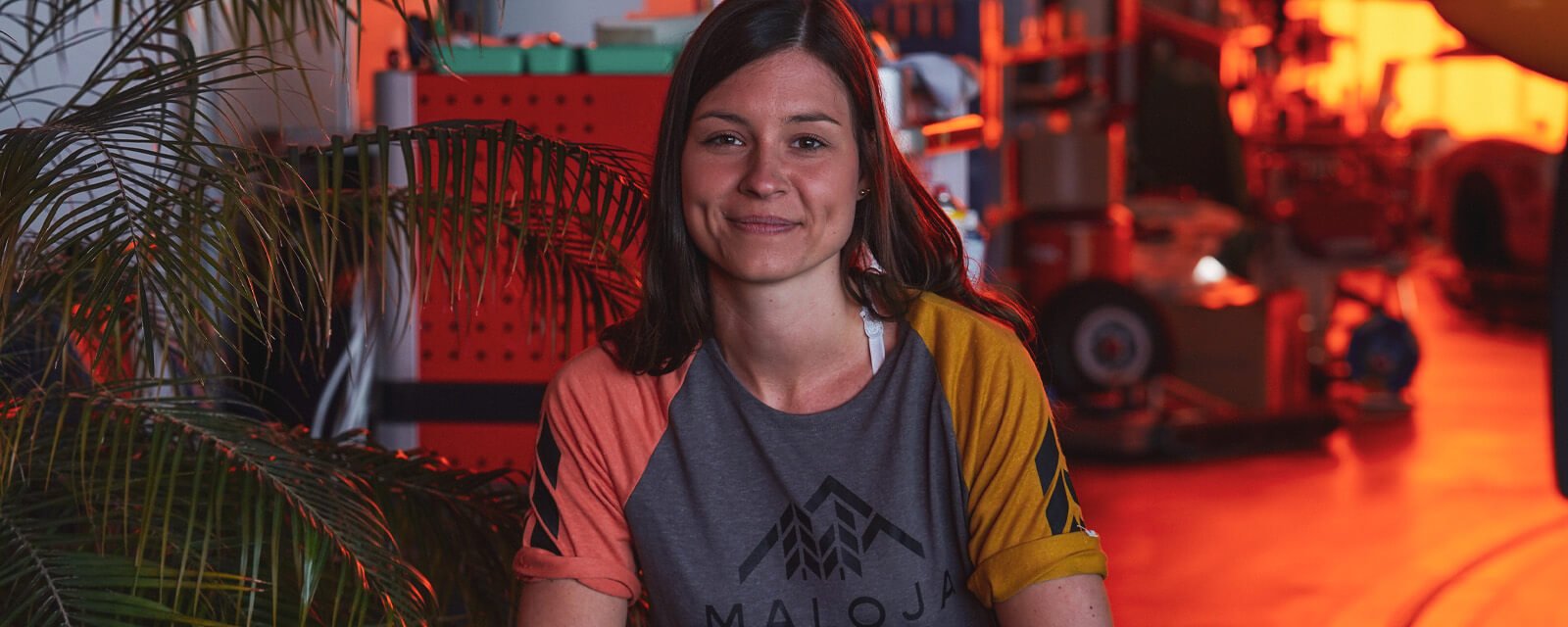 Person in einem Maloja-T-Shirt sitzt in einem industriellen Umfeld, umgeben von Pflanzen und Werkzeugen im Hintergrund.