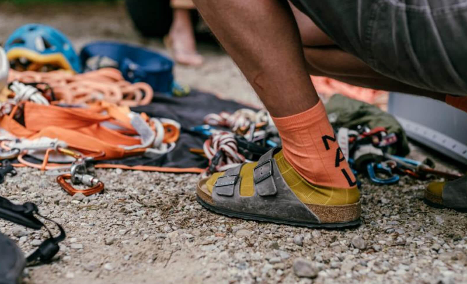Person in gelben Sandalen und orangen "Maloja"-Socken kniet neben Kletterausrüstung auf Schotterboden.