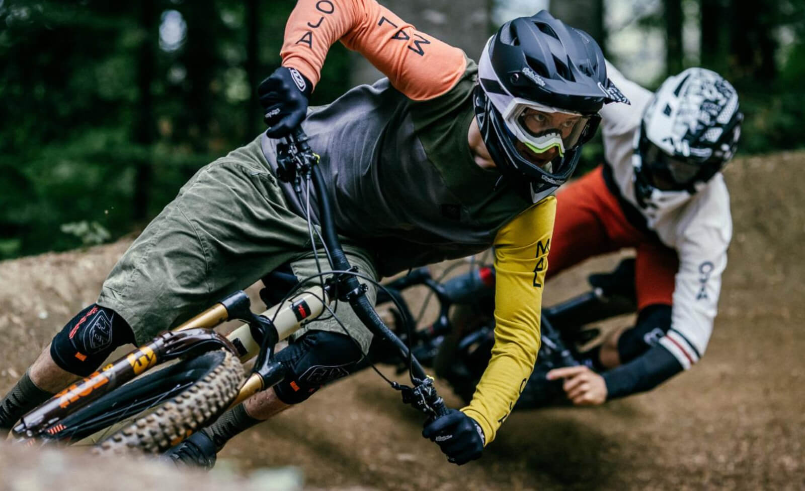 Mountainbiker in Maloja-Kleidung in einer Kurve auf Trail, Waldhintergrund, hart am Limit fahrend.