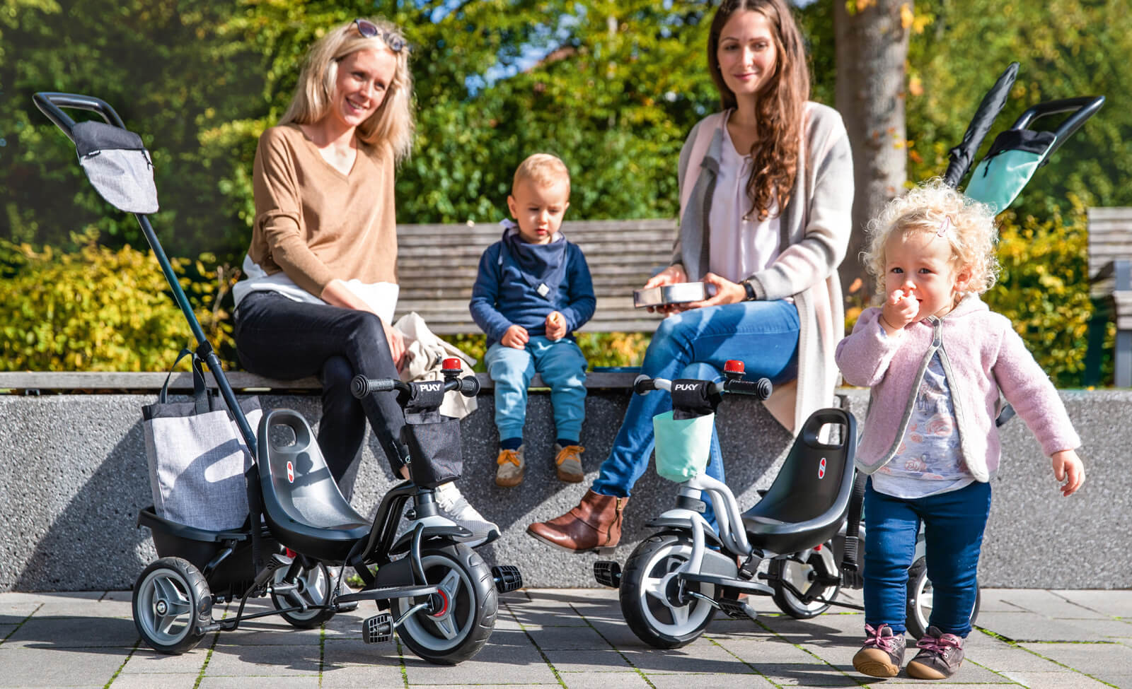 Zwei Kinder auf Puky-Dreirädern mit Erwachsenen auf einer Bank im Park. Tragegriffe und kleines Kind im Vordergrund.