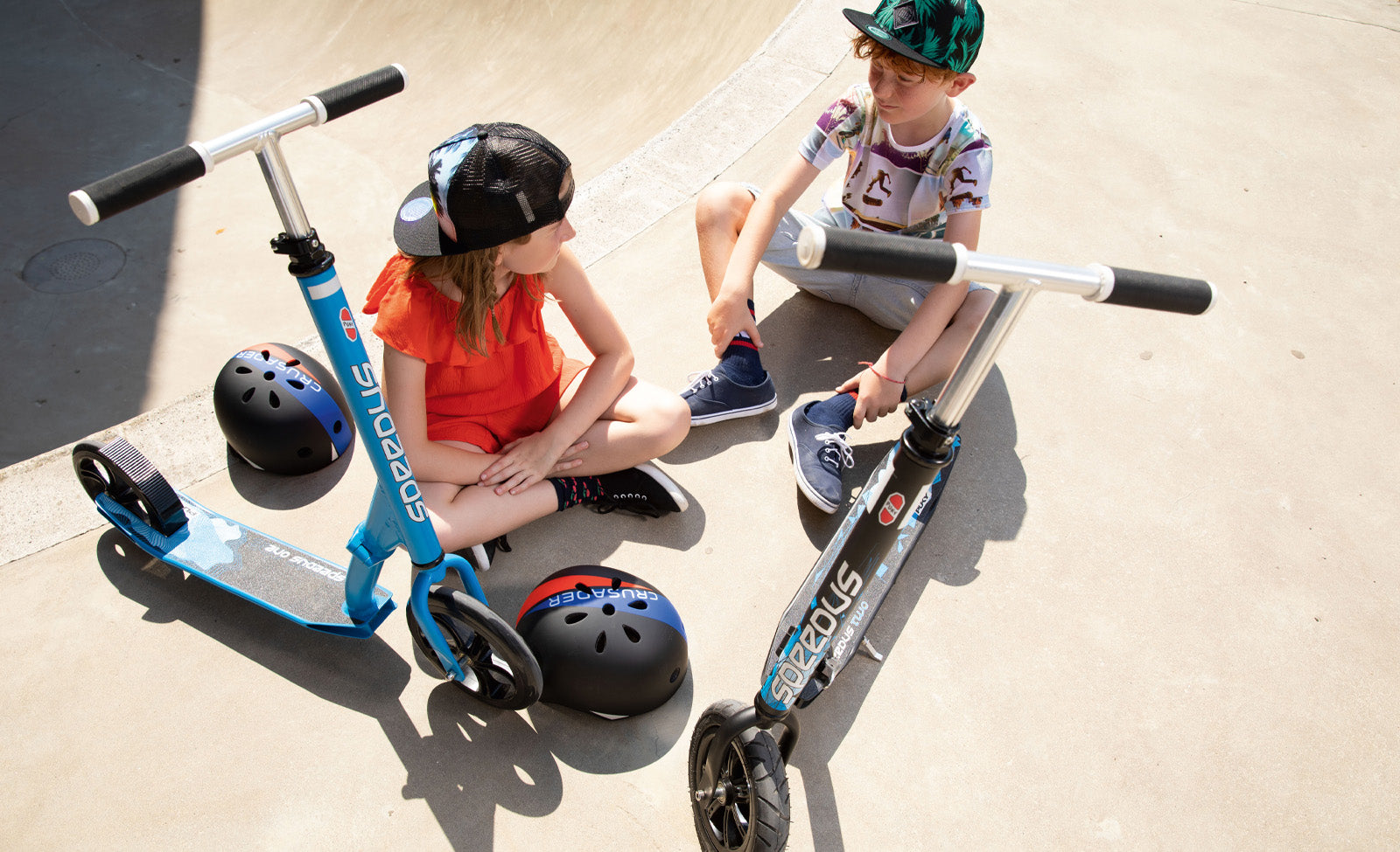 Zwei Kinder mit Scooter-Sporthelmen sitzen neben blauen und grauen Kinderrollern der Marke Scoot&Ride.