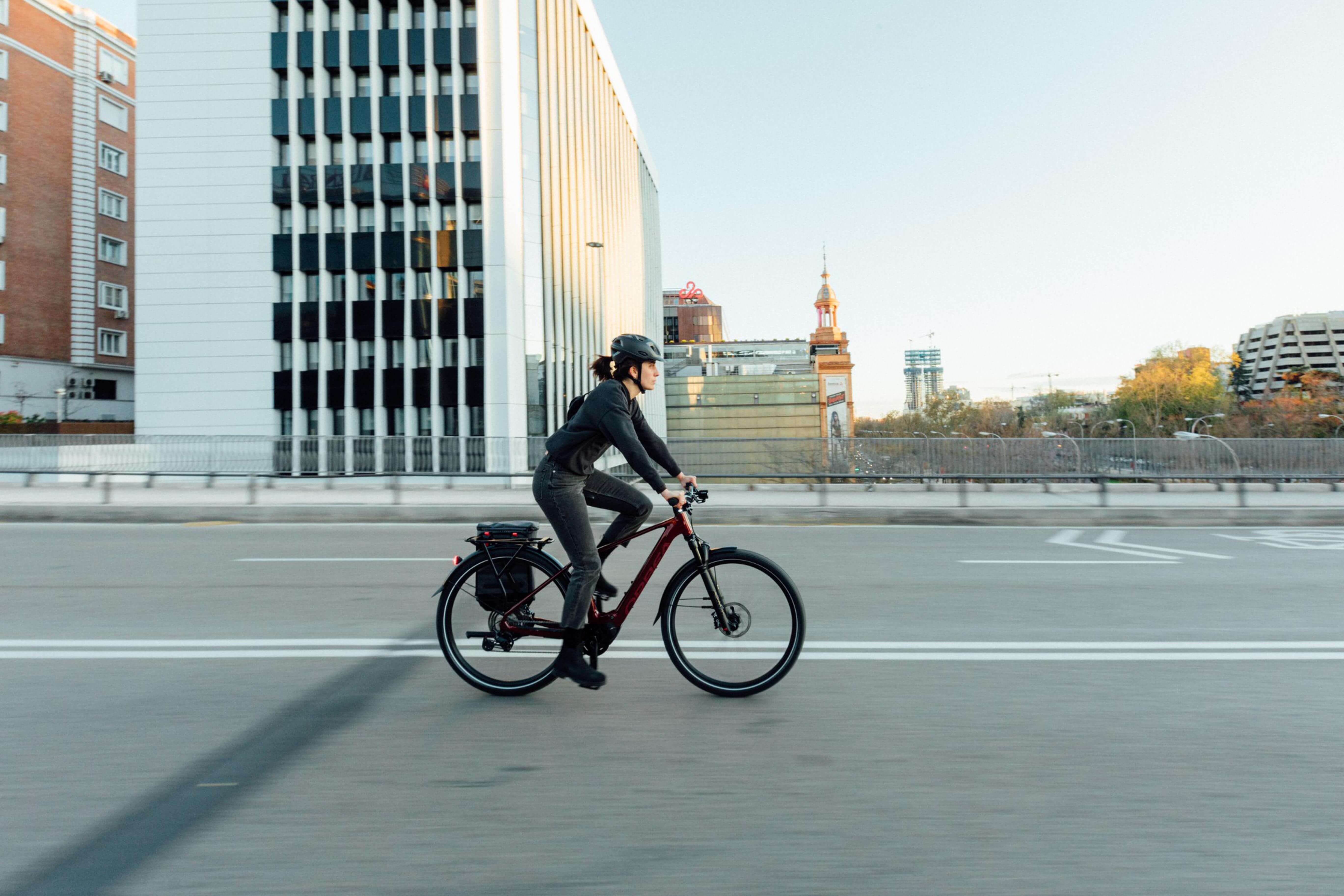 Person fährt Orbea-Elektro-Trekkingrad in der Stadt, trägt Helm, modernes Bürogebäude im Hintergrund.