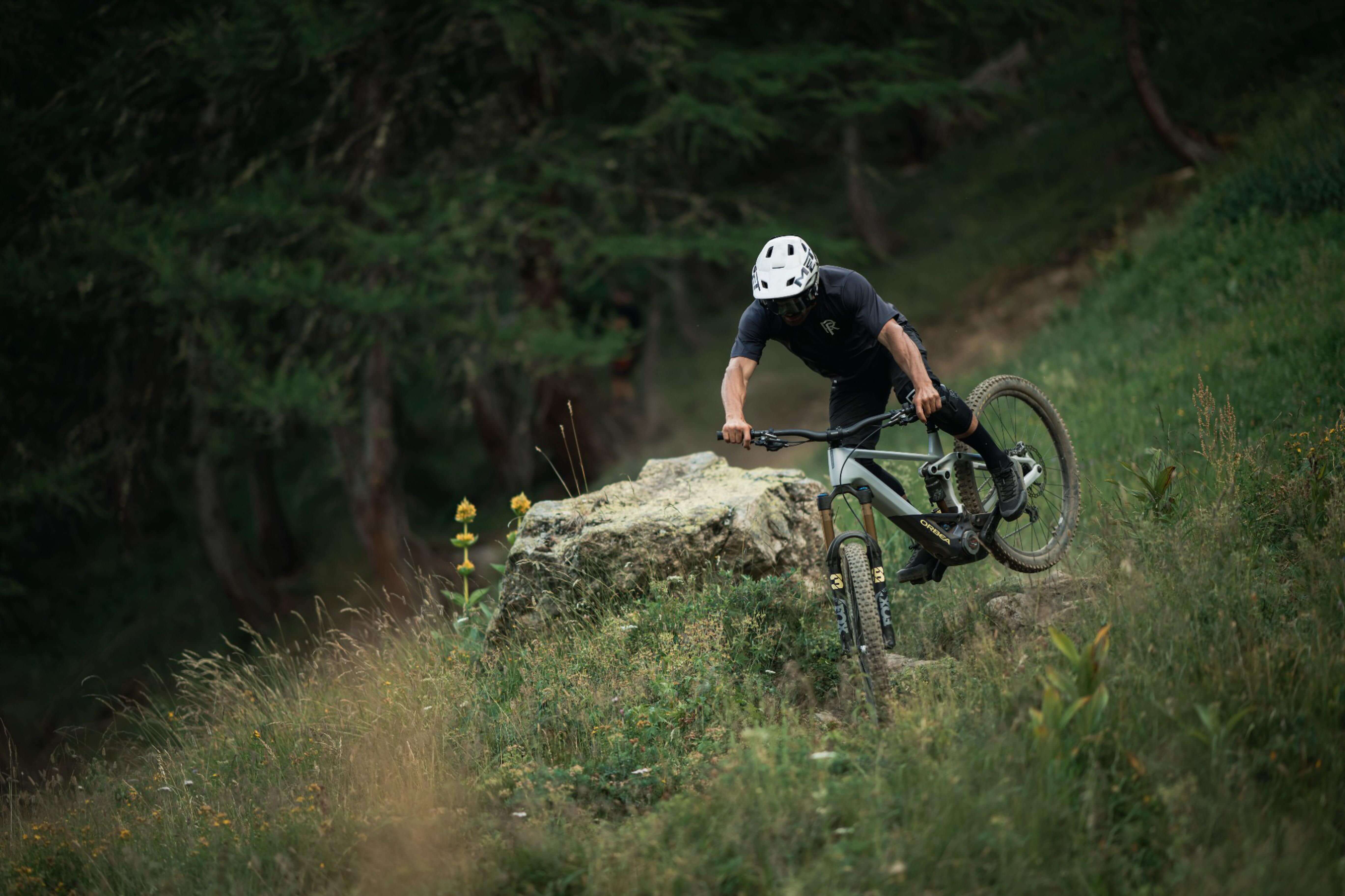 Mountainbiker in schwarzer Ausrüstung fährt ein Orbea E-MTB über steinigen Waldweg. Modell nicht sichtbar.