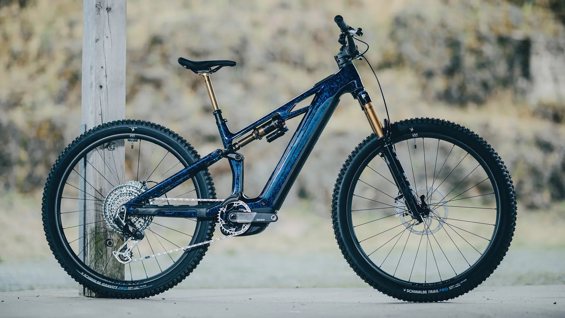 Blaues CUBE STEREO HYBRID ONE44 HPC SLT 800 mit Vollfederung, geeignet für den Geländeeinsatz, abgestellt an einem Holzbalken. Schwalbe-Reifen.