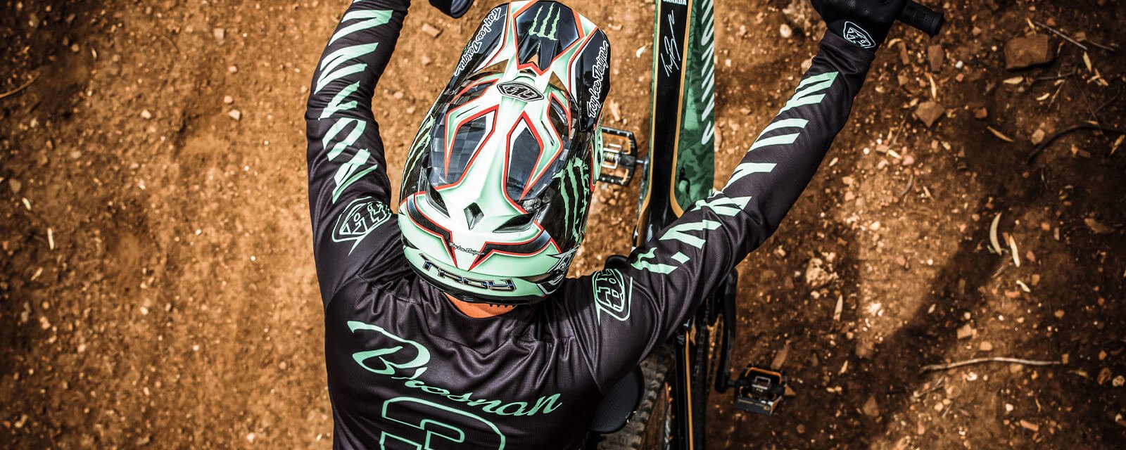 Biker mit Troy Lee Designs Helm auf Mountainbike auf staubigem Pfad, Rückansicht, markante Helmgestaltung.