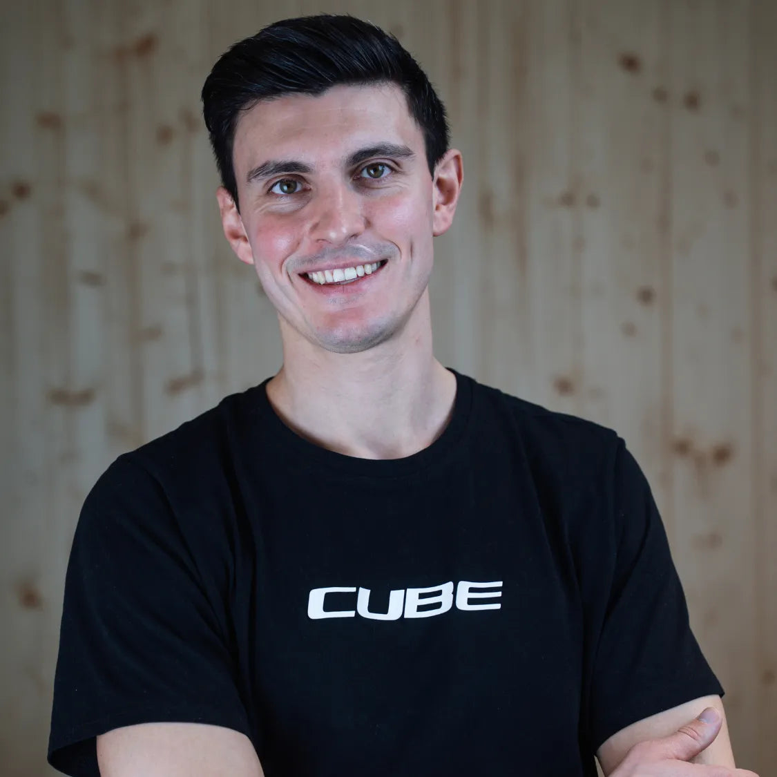 Porträt eines lächelnden Mannes in schwarzem T-Shirt mit weißem Cube-Logo vor Holzwand - Dima, Ansprechpartner in Paderborn.