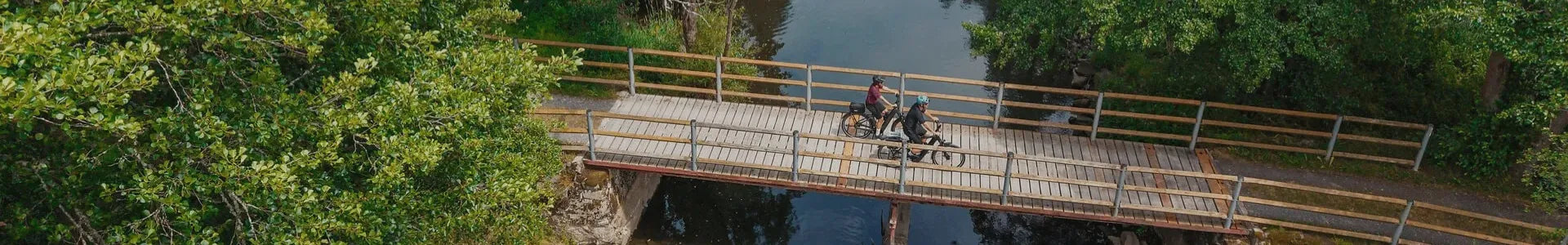 Zwei Personen radeln auf einer Holzbrücke über einen Fluss, umgeben von grüner Natur. Cube Kathmandu Hybrid 2026 Ideal für Outdoor-Aktivitäten.