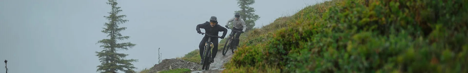 Zwei Mountainbiker fahren auf einem schmalen, matschigen Pfad bergab zwischen Bäumen und Sträuchern bei nebligem Wetter.
