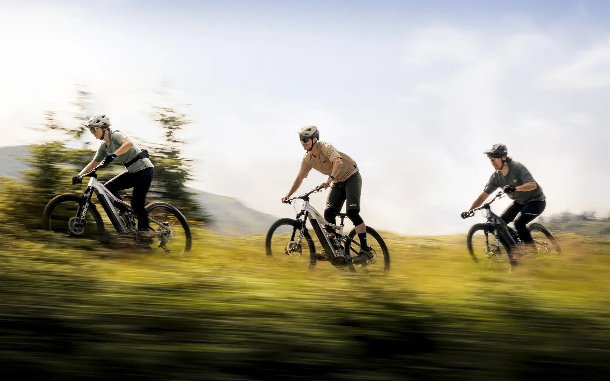 Drei Mountainbiker fahren mit E-Bikes schnell durch eine grüne Landschaft. Atmosphäre ist sportlich und dynamisch.