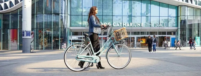 Frau schiebt pastellgrünes Gazelle Citybike mit Holzkorb vor Einkaufszentrum, modernes städtisches Pendlerfahrrad.