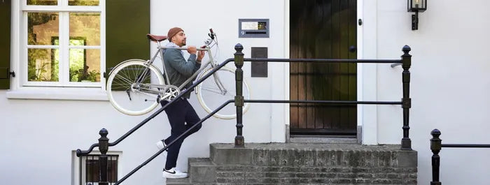 Person trägt ein silberfarbenes Gazelle Fahrrad die Treppe eines weißen Hauses hoch. Ideal für urbane Mobilität.