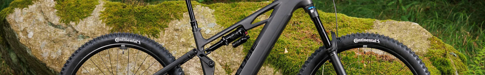 Schwarzes Mountainbike Cube Stereo Hybrid One77 im Gelände, mit Continental-Reifen, vor einem mit Moos bedeckten Felsen platziert.