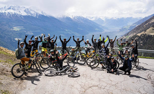 Gruppe von Mountainbikern posiert auf Bergstraße mit Blick auf verschneite Berge.