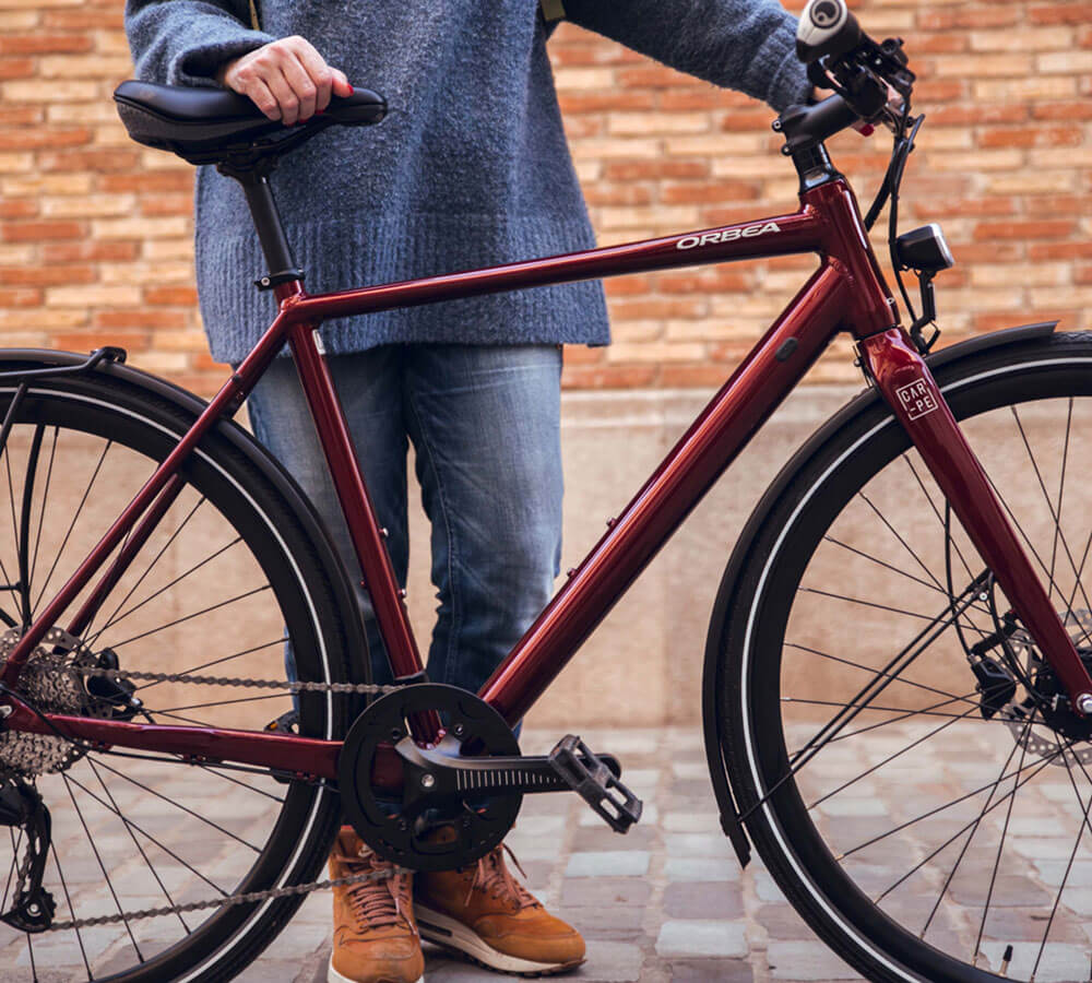 Rotes Orbea Citybike, Modell Carpe, für den urbanen Gebrauch, mit aufrechtem Fahrer in Jeans und Stiefeln.