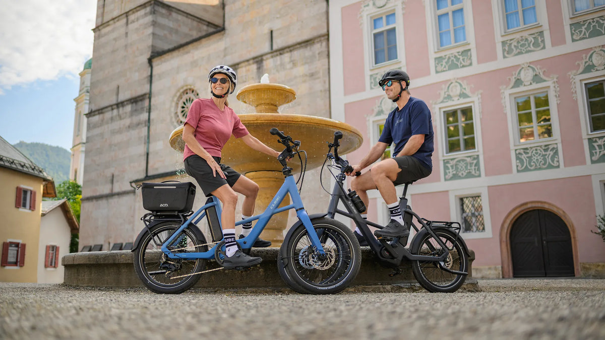 Zwei Personen auf CUBE COMPACT HYBRID DELUXE 540 vor einem Brunnen, tragen Helme und Sportkleidung, entspannte Stadtszene im Hintergrund.